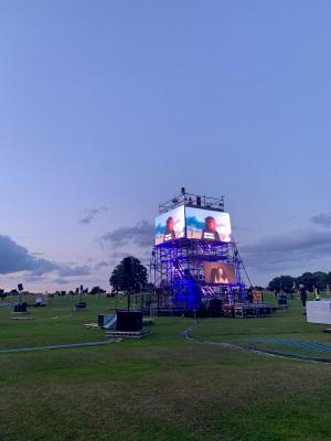 Performance of 'Hear Us!' on a large scaffolding tower in park with platform at the top where young peopel went to make speeches and sing. They are projected on large screens, lit up against the fading light